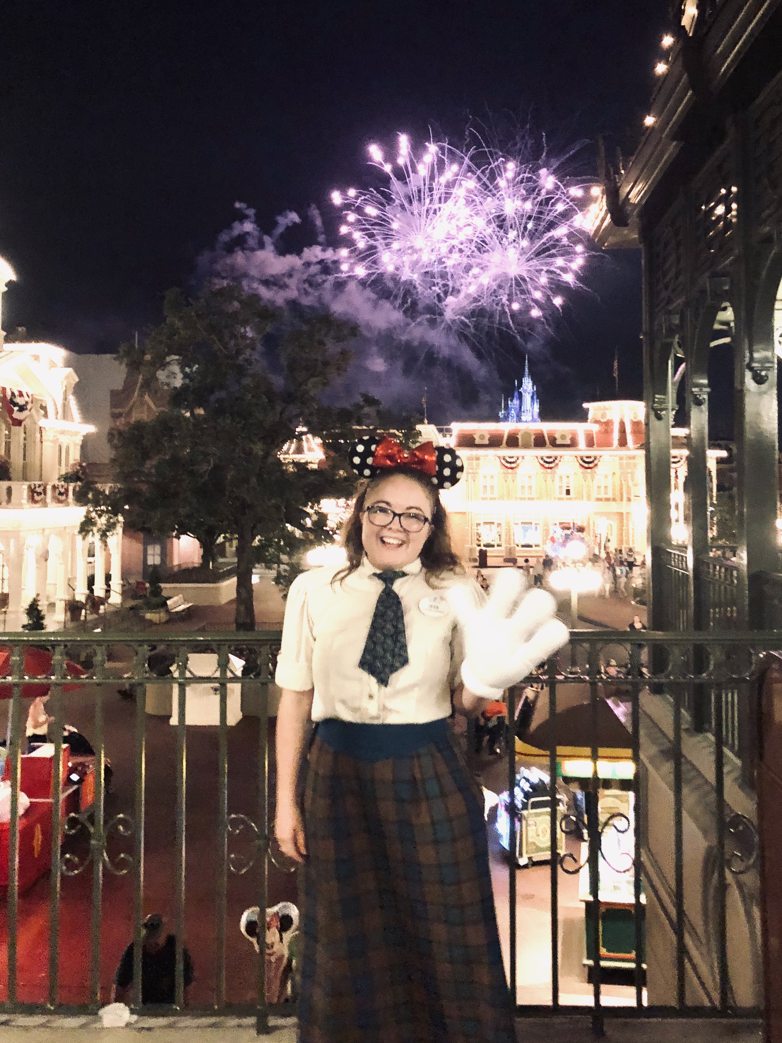 Disney cast member dressed in emporium plaid costume waving with Mickey glove with Main Street USA and fireworks in the background