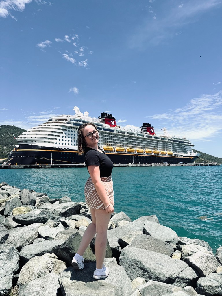 Girl standing on rocks turning and looking back at camera with ocean and Disney cruise ship in the background in the Virgin Islands