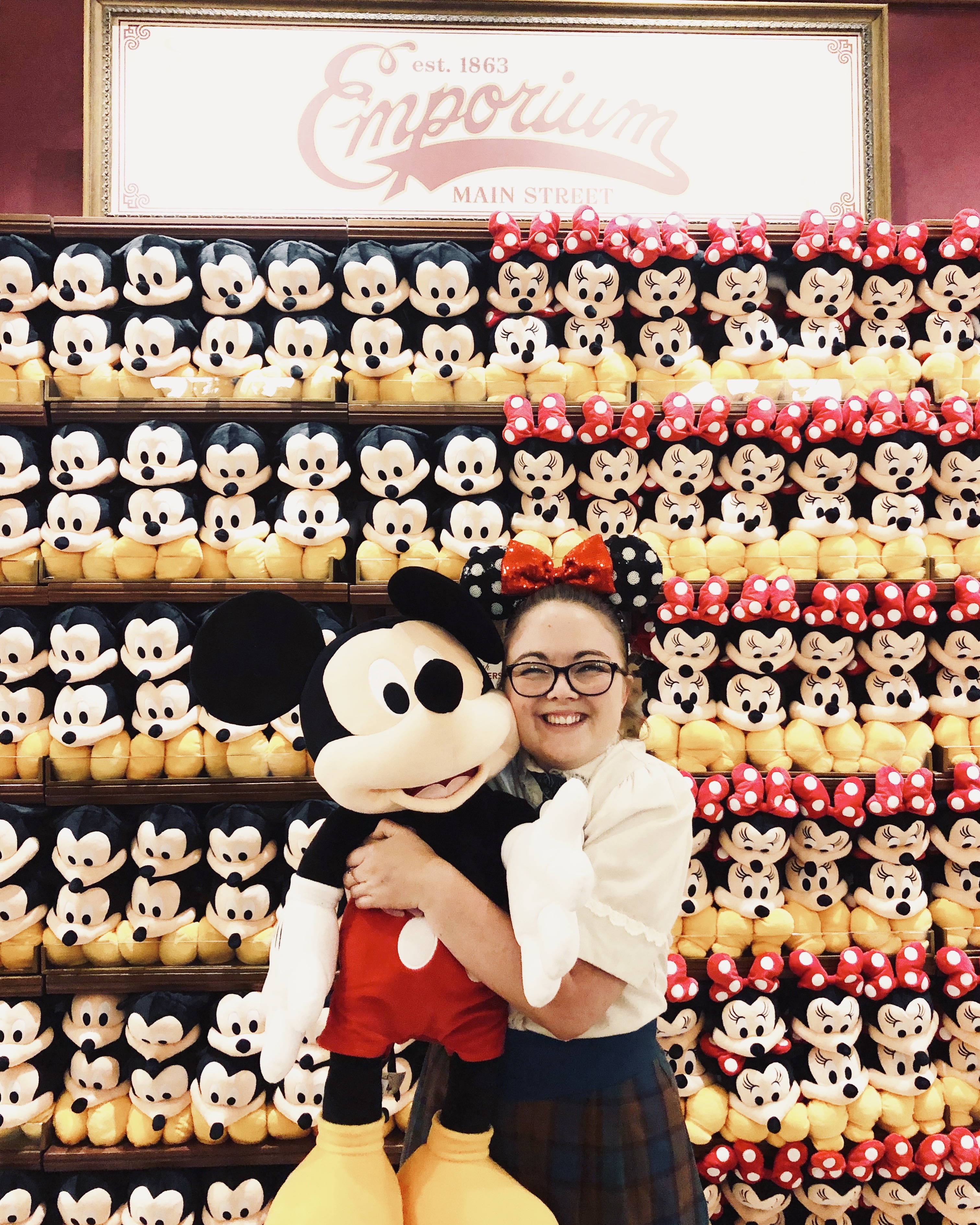 Disney cast member dressed in plaid costume standing in-front of Mickey plush wall in the emporium while hugging a Mickey plush