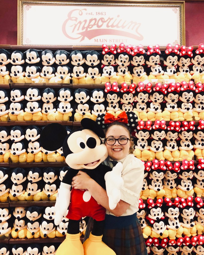 Disney cast member dressed in plaid costume standing in-front of Mickey plush wall in the emporium while hugging a Mickey plush