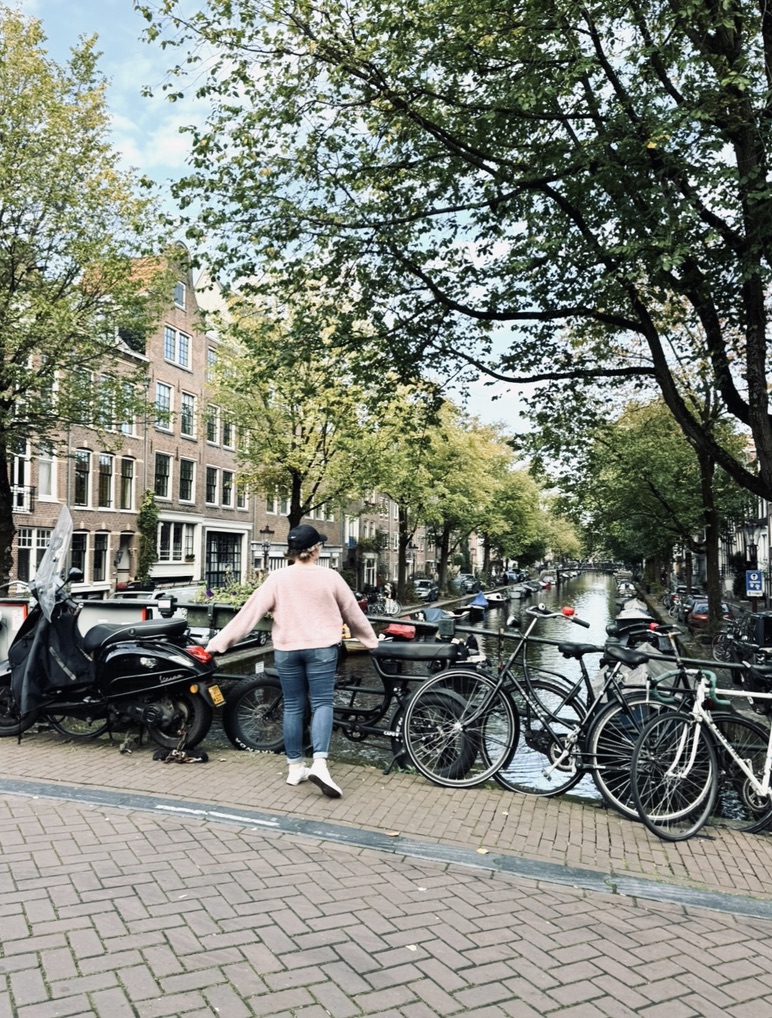 Girl walking towards railing with a background of colorful houses along a canal with bicycles and green trees in Amsterdam, The Netherlands