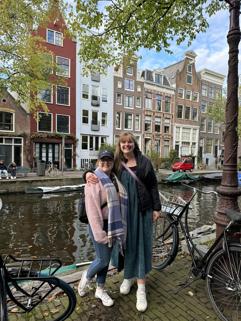 Two friends standing in front of colorful houses along a canal with bicycles and green trees in Amsterdam, The Netherlands