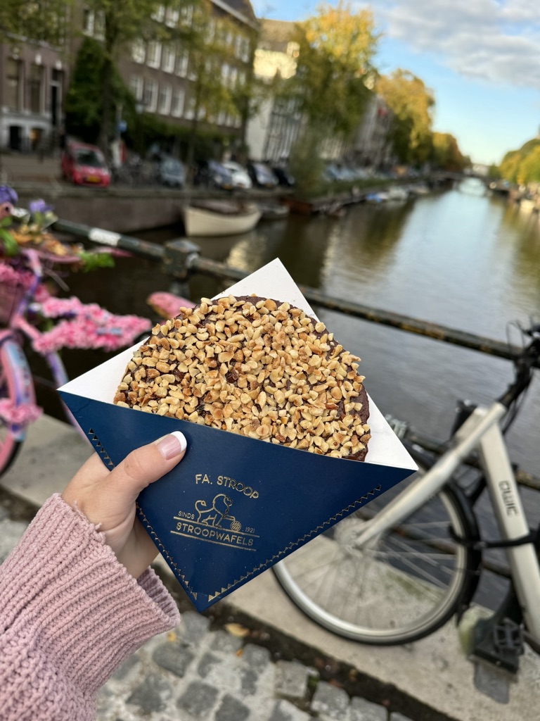 Hand holding stroowafel in front of colorful houses along a canal with bicycles and green trees in Amsterdam, The Netherlands