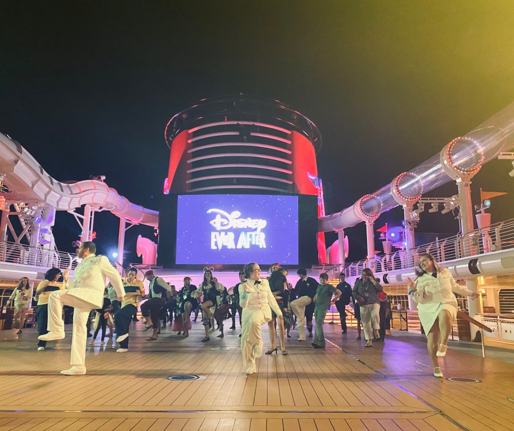 Disney Cruise Line ship open deck at night with a group of people dancing. Three ship officers in the front teaching a dance routine.