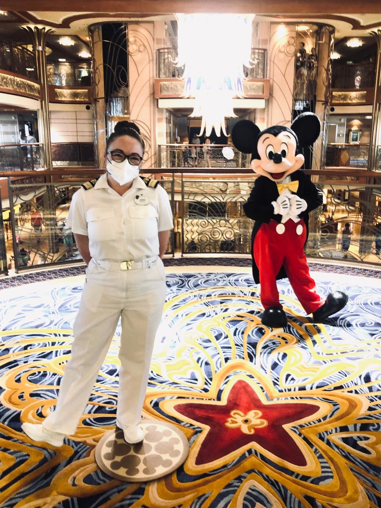Disney cruise ship officer wearing a face mask posing with Mickey Mouse in atrium on Disney Dream cruise ship