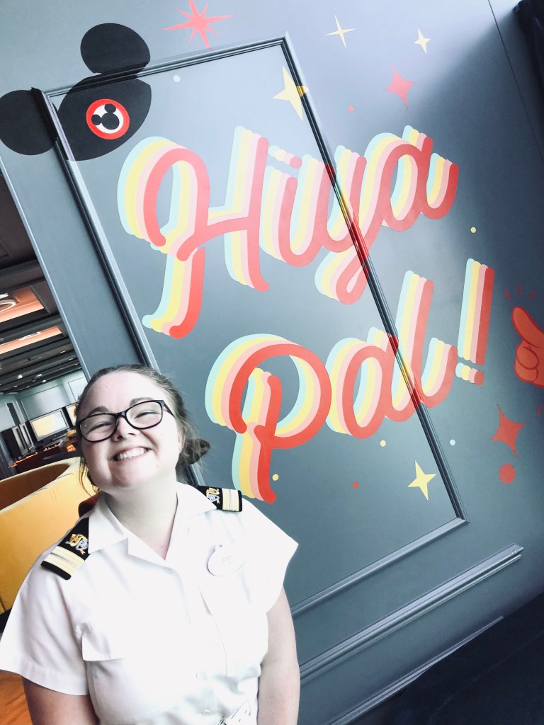 Disney cruise line officer smiling in Vibe kids club in front of 'Hiya Pal' artwork on the Disney Wish
