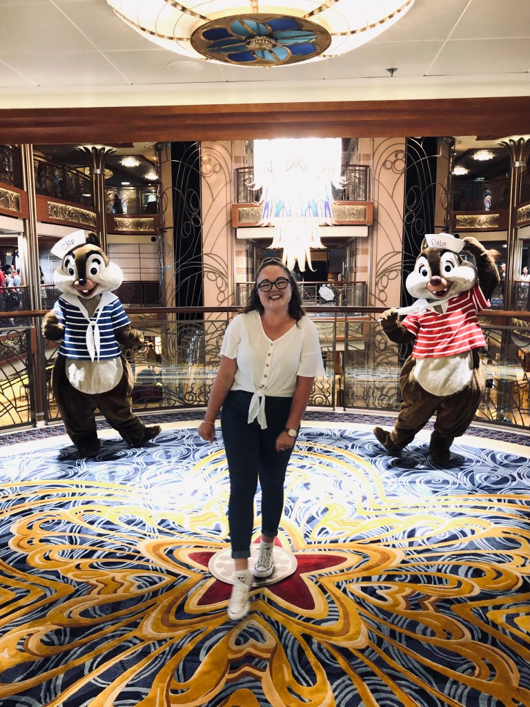 Girl smiling with Chip and Dale onboard the Disney Dream with atrium chandelier in background.