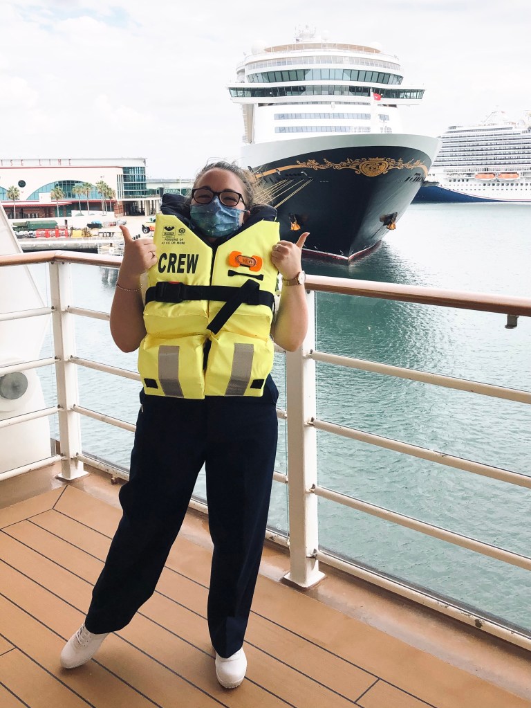 Disney Crew Member wearing life jacket with thumbs up while standing on deck with Disney Cruise ship in the background.