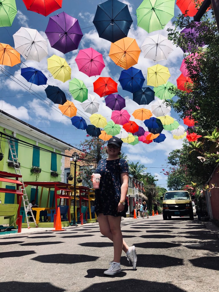 Girl standing on street with a starbucks pink drink on a street with colorful umbrellas hanging above in Nassau, Bahamas