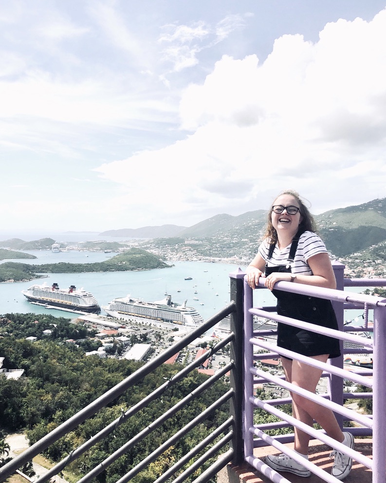Girl smiling while holding railing with view of St Thomas from Paradise Point observation deck in the distance. Ocean, islands and ship on a sunny day.