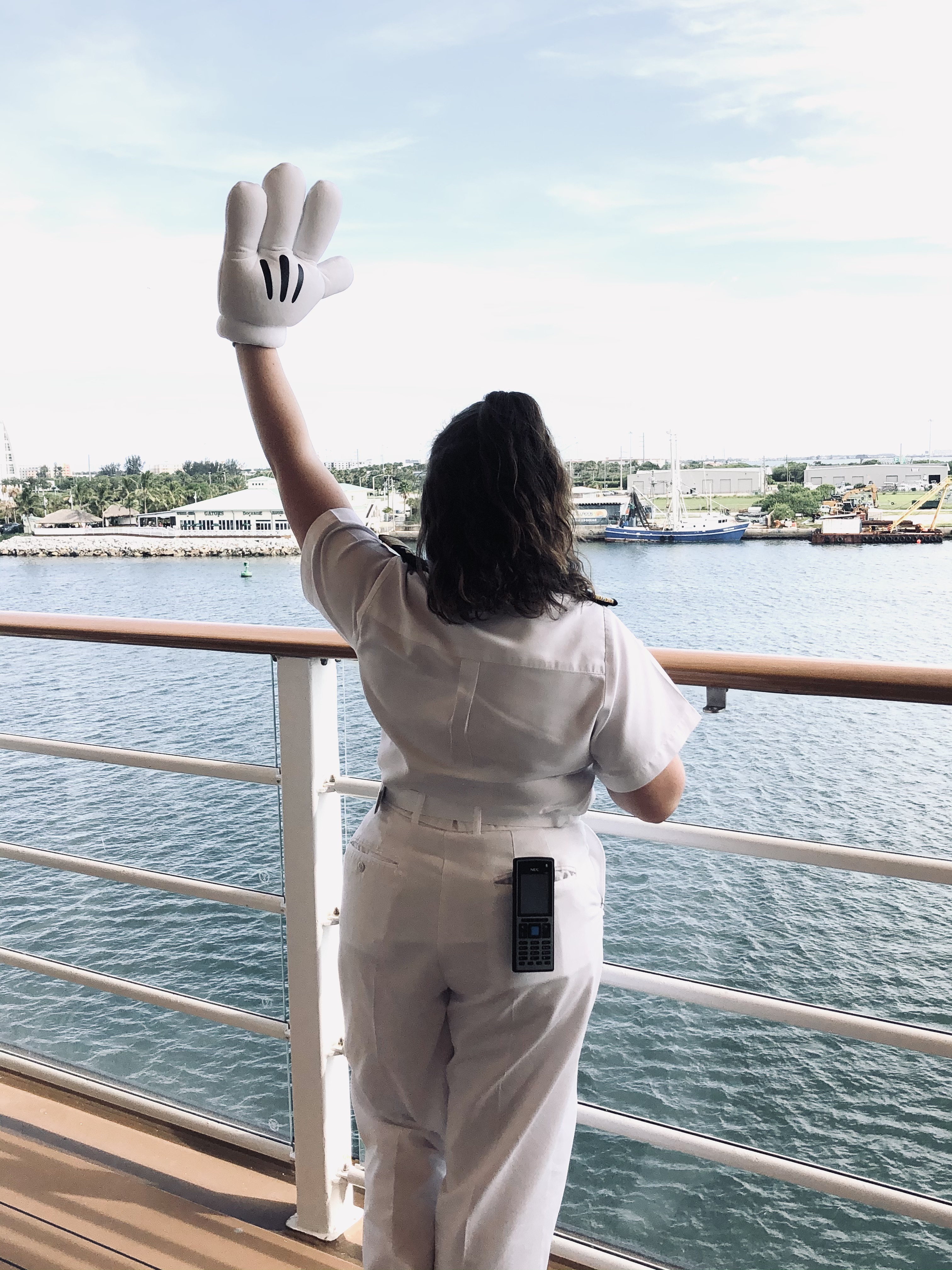 Disney Cruise Line officer waving with a mickey glove while standing on deck with ocean and Port Canaveral in the background.