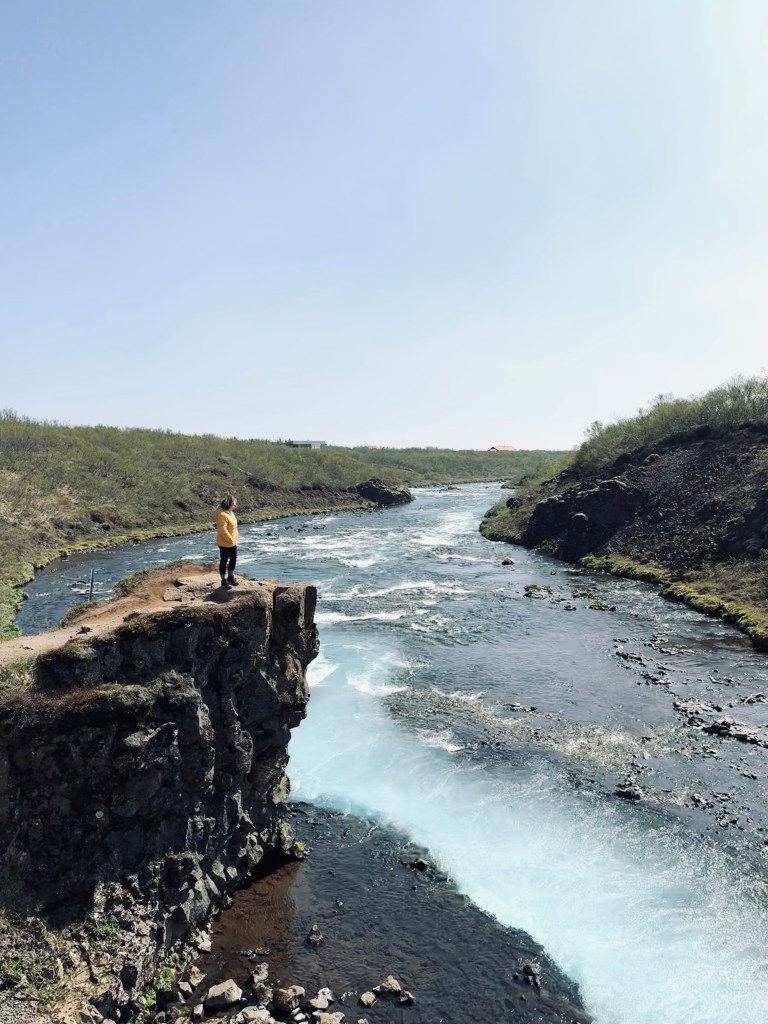 Girl in yellow coat standing on cliff looking over Bruarfoss waterfall in Iceland