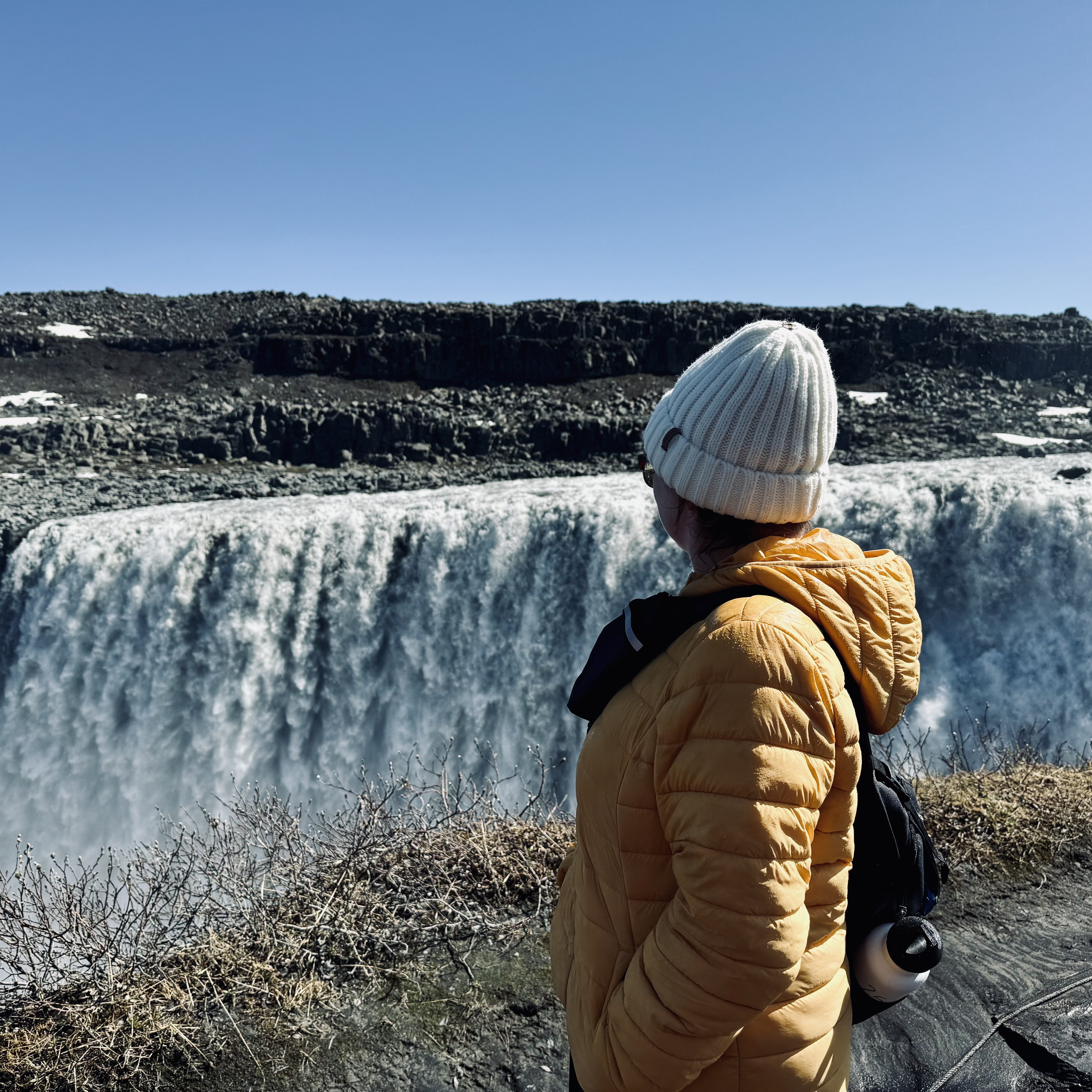 Girl in yellow coat looking out over rushing waterfall at Dettifoss, Iceland