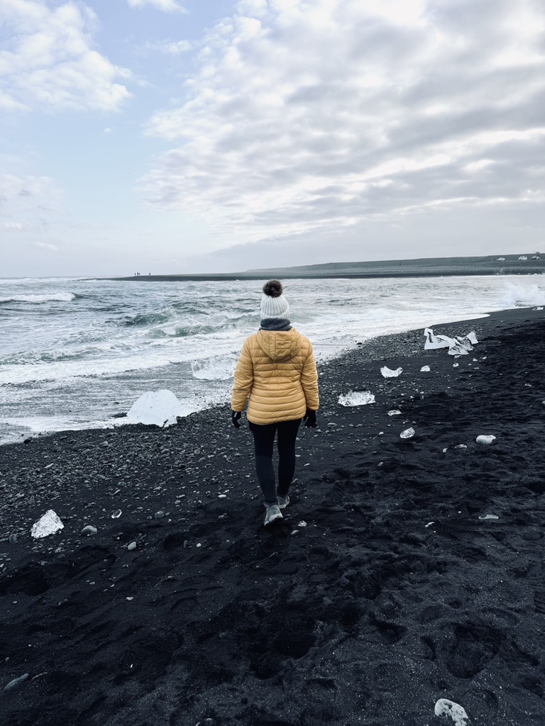 Girl in yellow coat walking towards ocean on black sand beach surrounded by ice chunks at Diamond Beach, Iceland