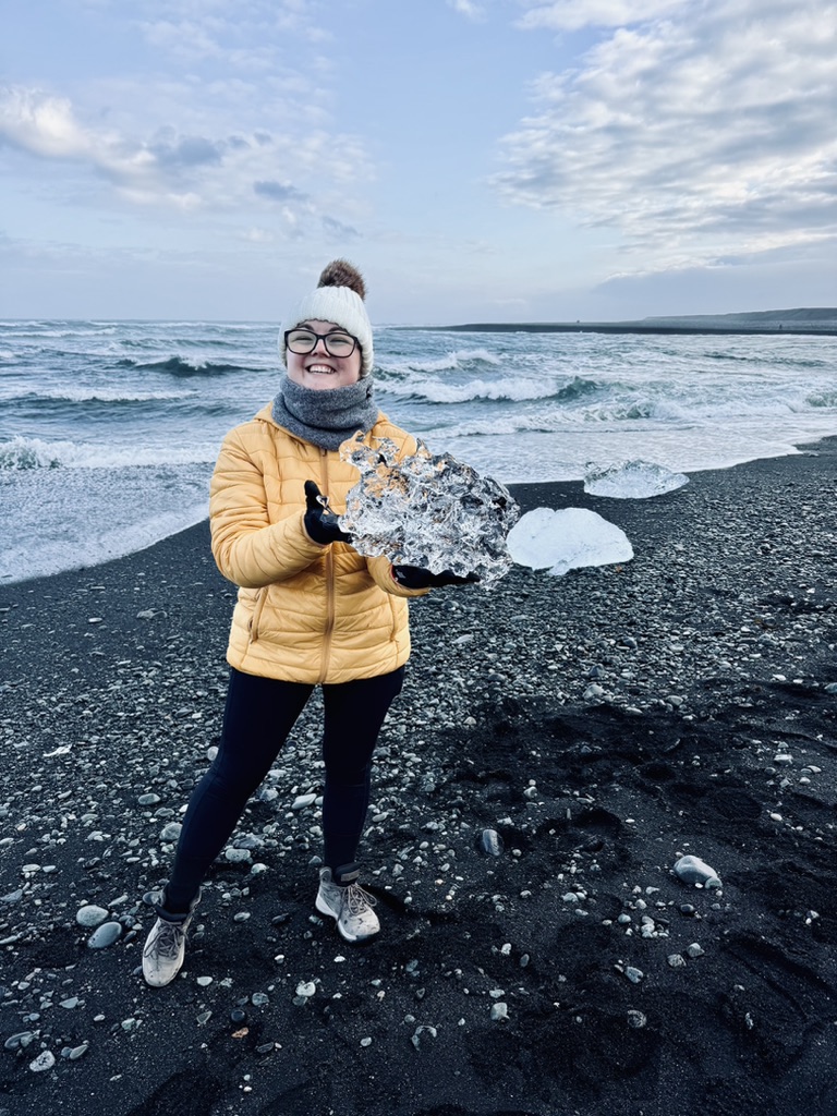 Girl in yellow coat holding a big chunk of ice that washed up on black sand beach at Diamond Beach, Iceland