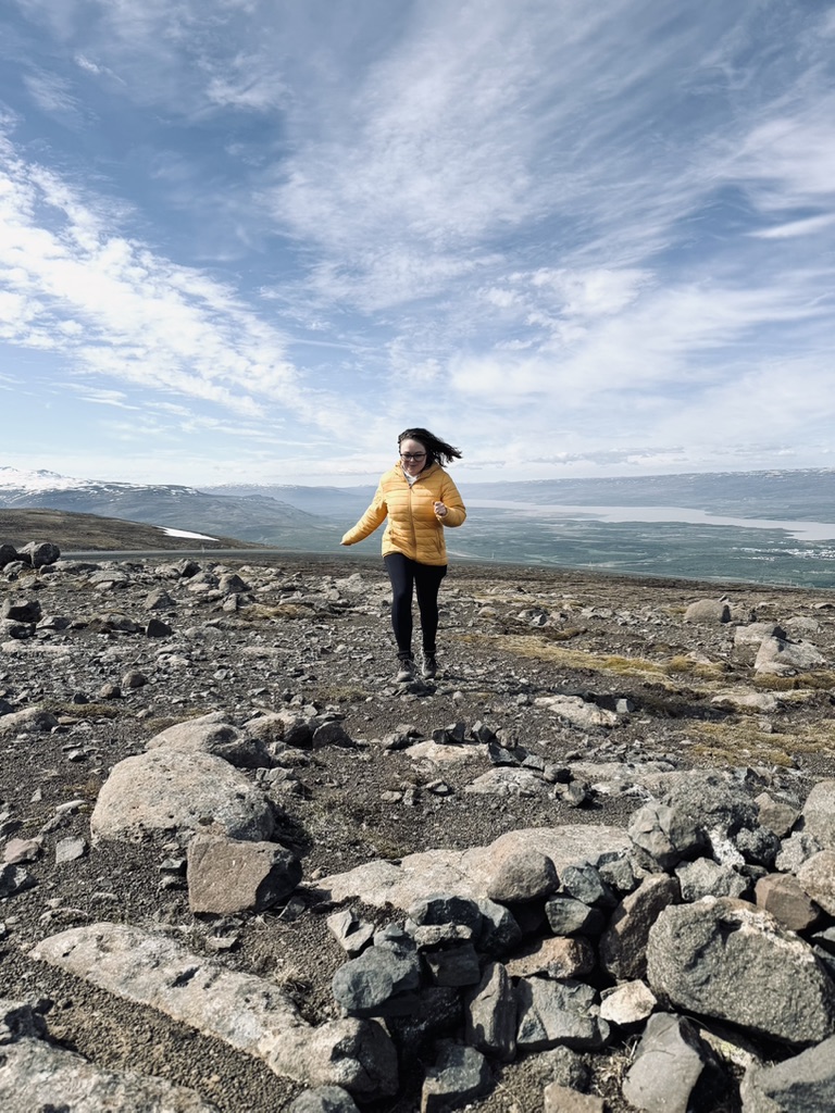 Girl in yellow coat running on rocks on the top of a hill with valley in the background in East Iceland