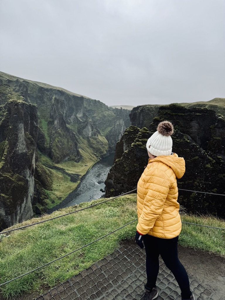 Girl in yellow coat looking out at deep, green canyon at Fjaðrárgljúfur Canyon, Iceland