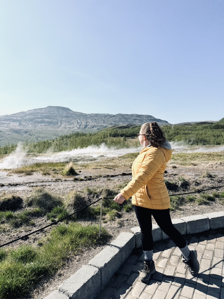 Girl in yellow coat looking out over steaming pools at Geysir Geothermal Area, Iceland