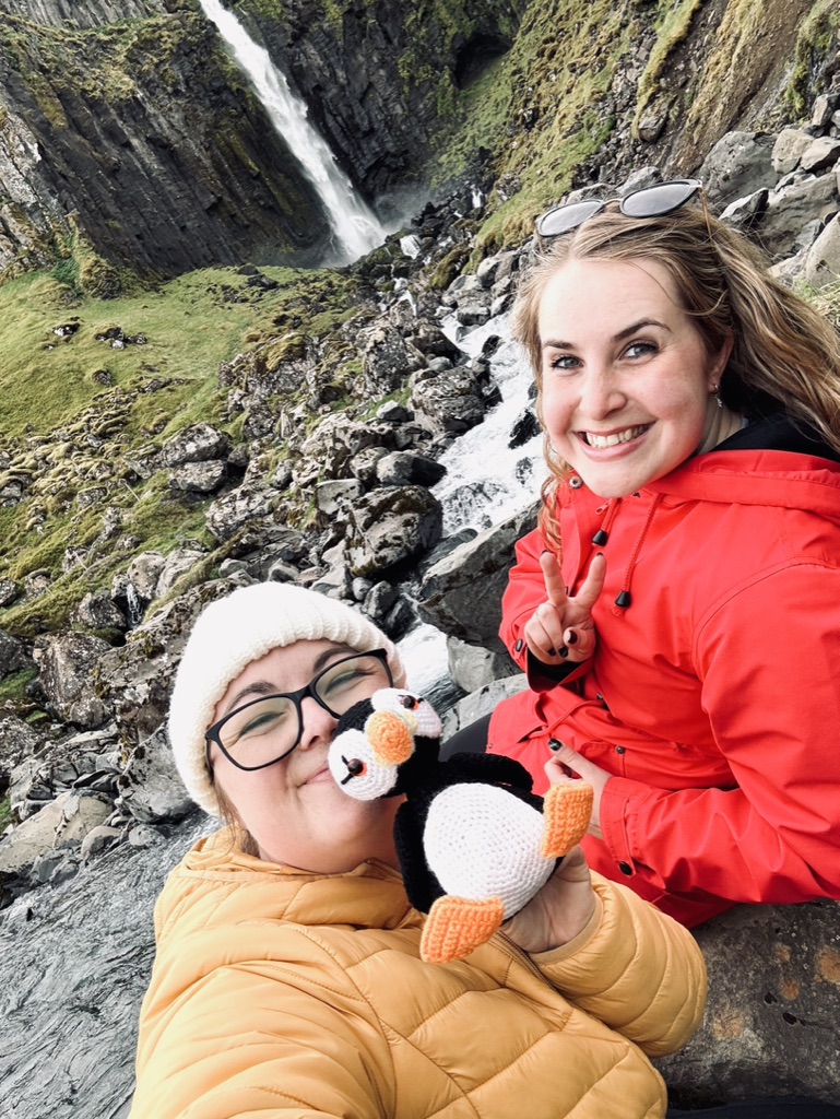 Two girls taking a selfie with a crochet puffin with Grundarfoss waterfall in the background, Iceland