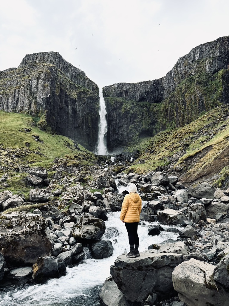 Girl in yellow jacket standing in front of Grundarfoss Waterfall in Iceland. Beautiful towering rocks and moss.