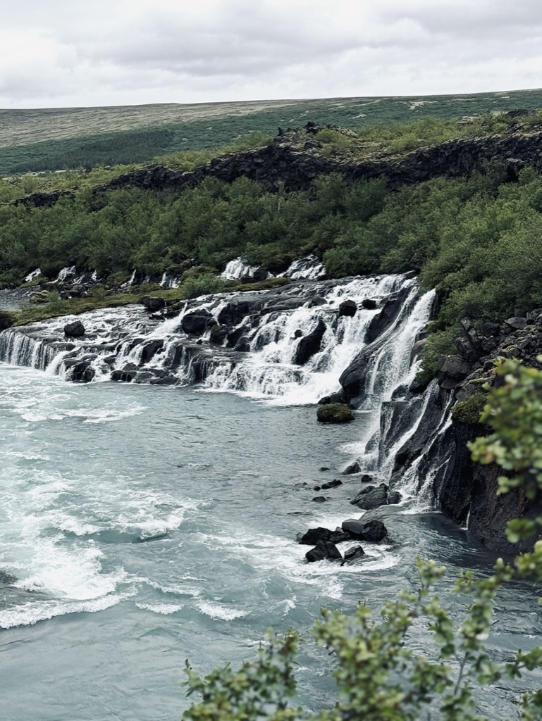Cascading waterfalls flowing from lava rocks with greenery at Hraunfossar Waterfall