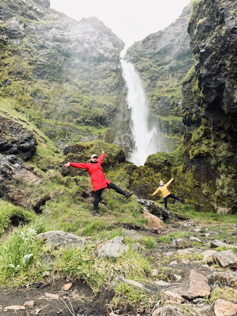 Two girls dancing in front of green mossy surrounds of Irafoss waterfall, Iceland