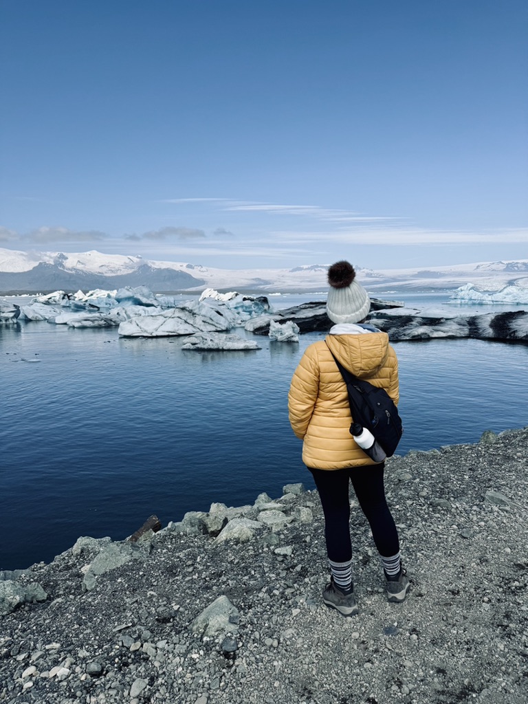 Girl in yellow coat looking out at icebergs floating on the blue waters of Jökulsárlón Glacier Lagoon, Iceland