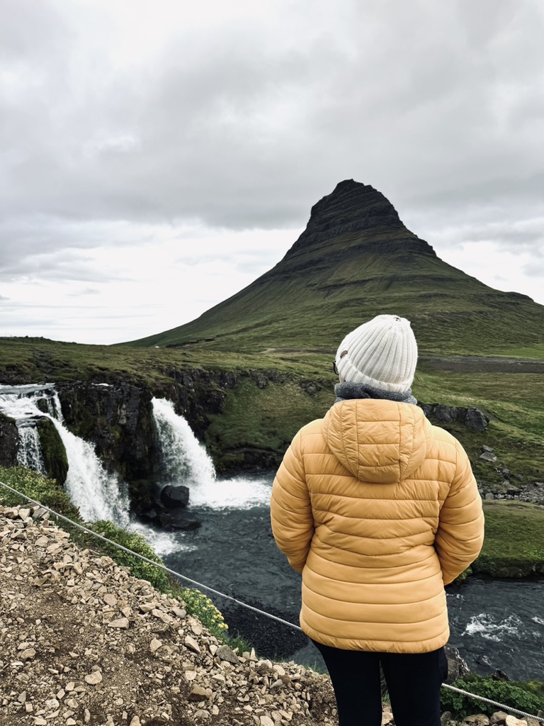 Girl in yellow coat looking out at green Kirkjufell Mountain in Iceland