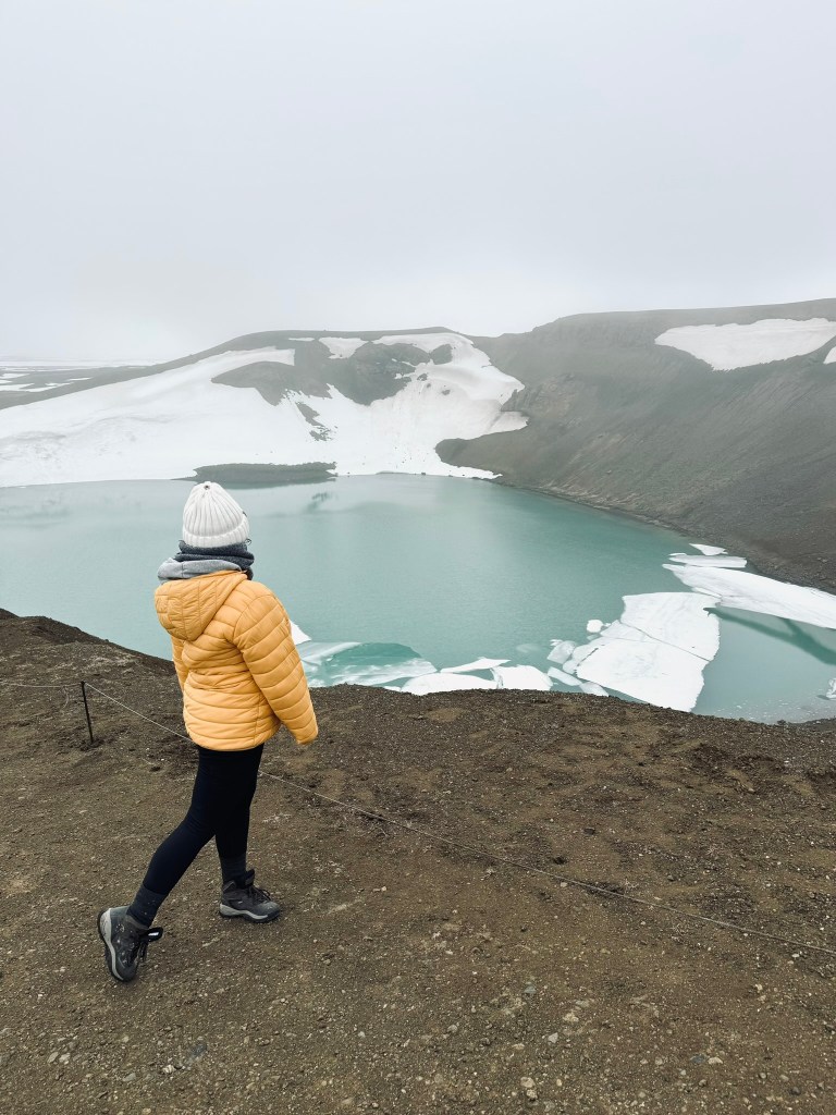 Girl in yellow coat looking out at blue water in crater around Mytavn, Iceland