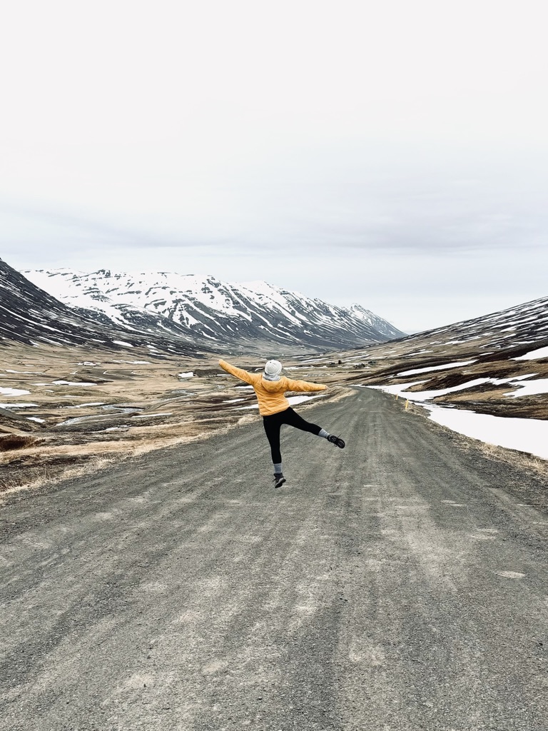 Girl in yellow coat jumping on empty road with snow capped mountains in distance in Iceland