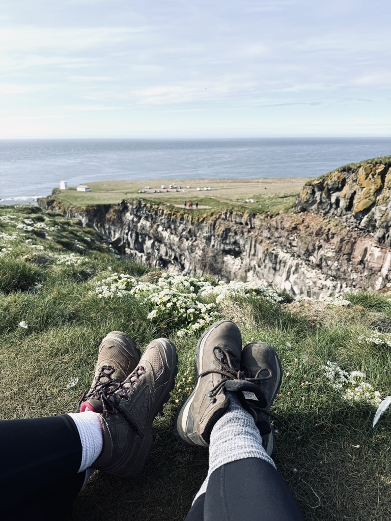 Two girls crossing feet with hiking boots with view of Puffin Cliffs in background in Westfjords, Iceland