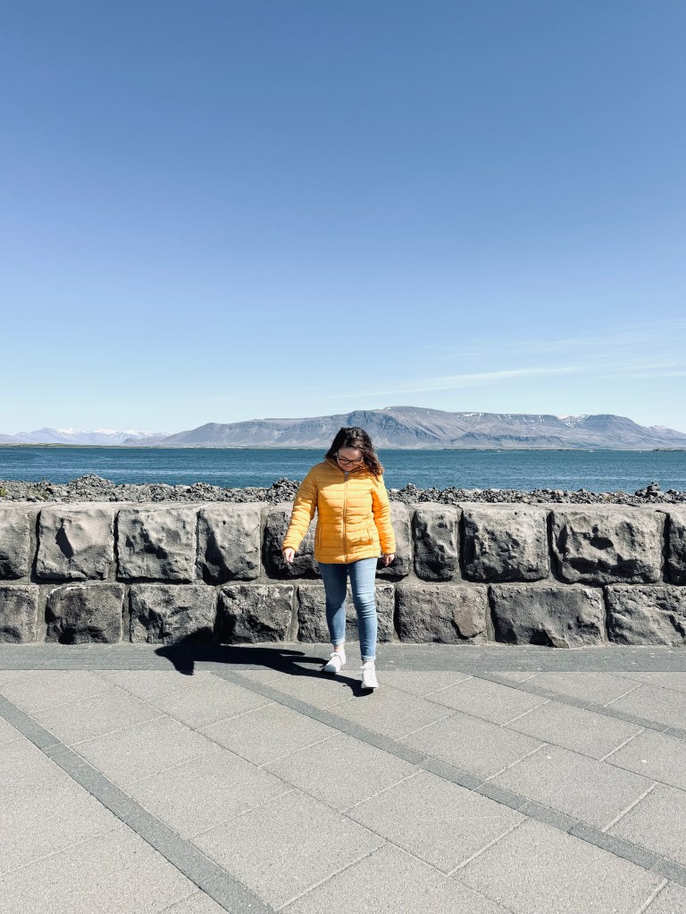 Girl in yellow coat walking on street with ocean, mountains and clear blue sky in background in Reykjavik, Iceland