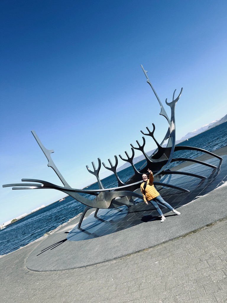 Girl in yellow coat posing beside Sun Voyager Monument in Reykjavik, Iceland on a sunny day