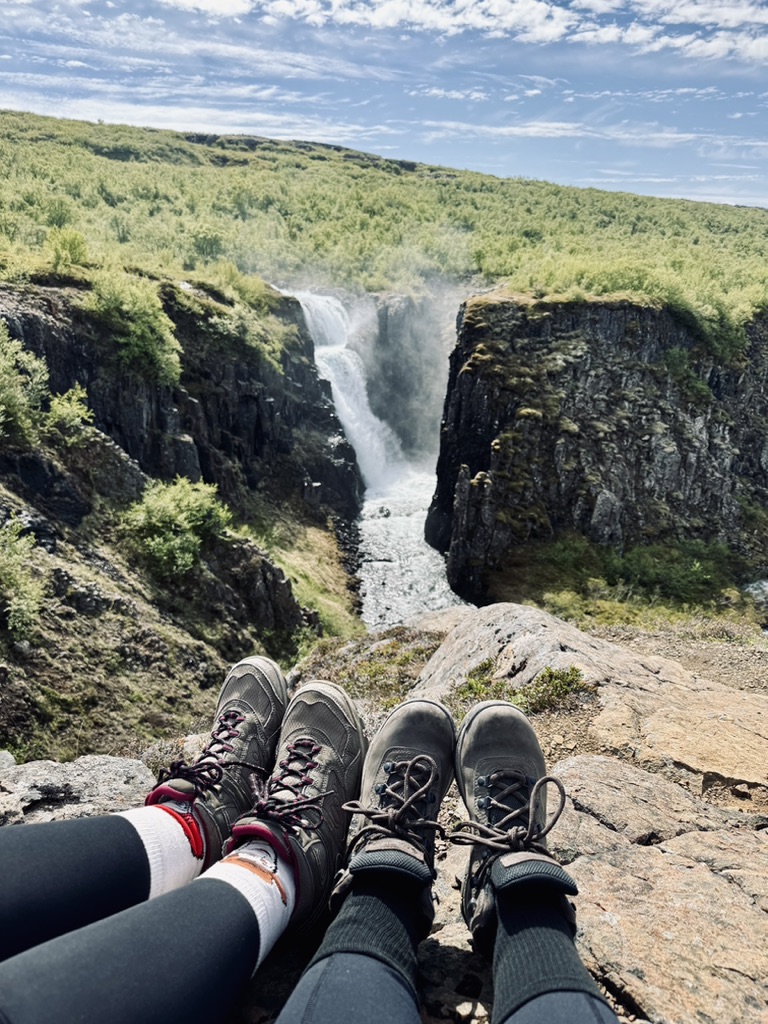 Two sets of hiking shoes having a hiking break with waterfall in the distance and green trees in Iceland