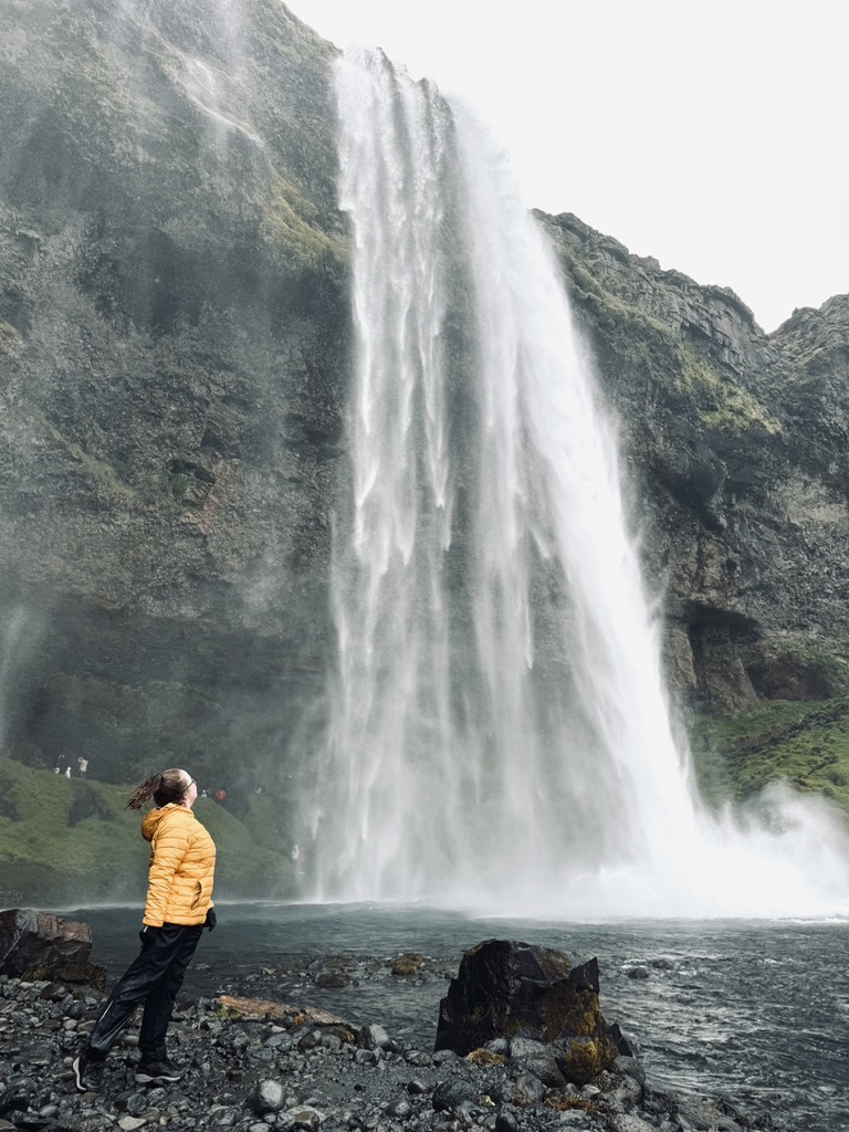 Girl in yellow coat looking up from the bottom of Seljalandafoss waterfall, Iceland