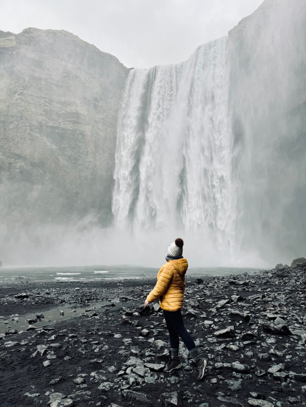 Girl in yellow jacket looking back at impressive Skogafoss waterfall in Iceland
