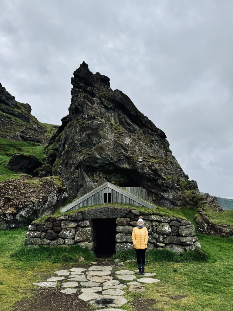 Girl in yellow coat smiling in front of rocky turf house in Iceland