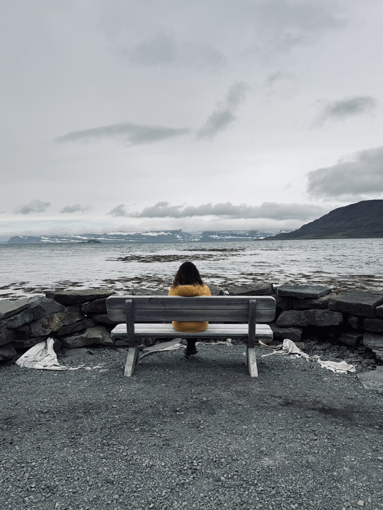 Girl sitting on bench looking out over ocean with mountains in distance in Iceland.