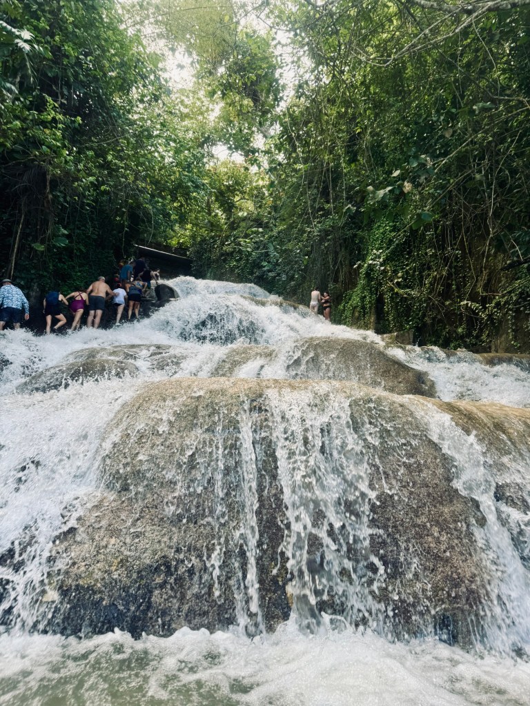 Waterfall flowing over large rocks with jungle background and people climbing up rocks at Dunns River Falls in Jamaica