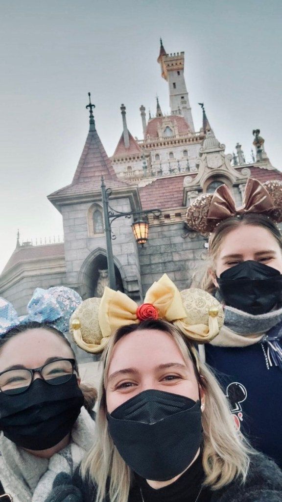 Three girls with minnie ears and facemasks taking a selfie in front of the Beauty and the Beast castle ride at Tokyo Disneyland.