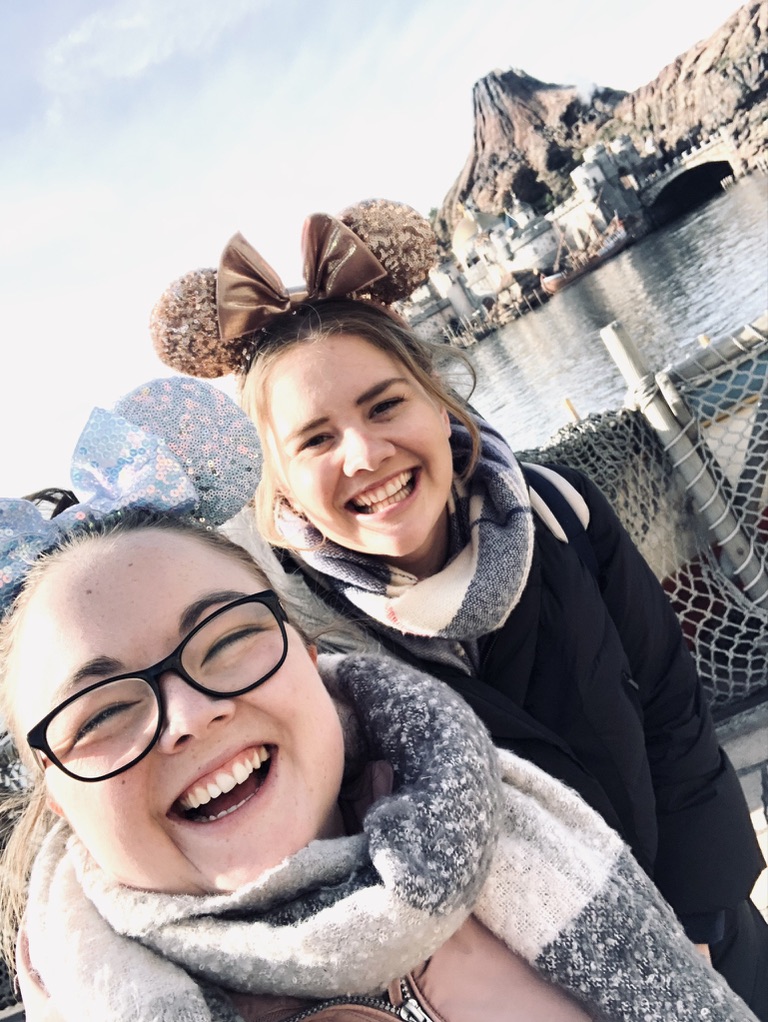 Two girls smiling wearing minnie mouse ears with Tokyo DisneySea lagoon and volcano in background.