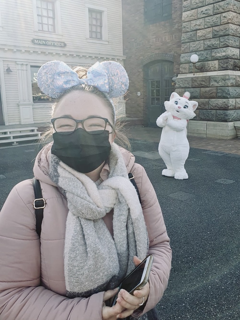 Girl smiling with facemask and minnie ears with Marie Disney Character in the background.