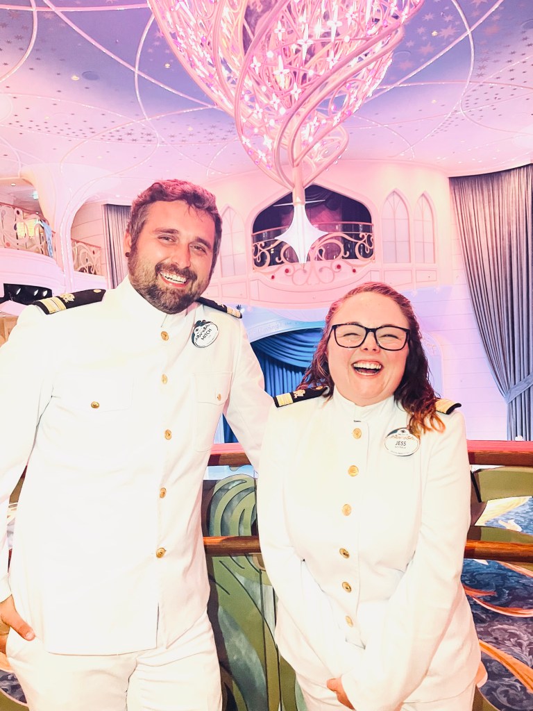 Two Disney Cruise Line officers smiling in Disney Wish Grand hall with chandelier in the background
