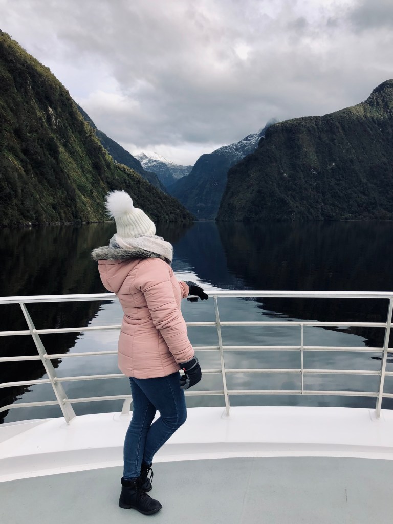 Girl in pink jacket on boat tour with green mountains and water reflection at Doubtful Sound, New Zealand
