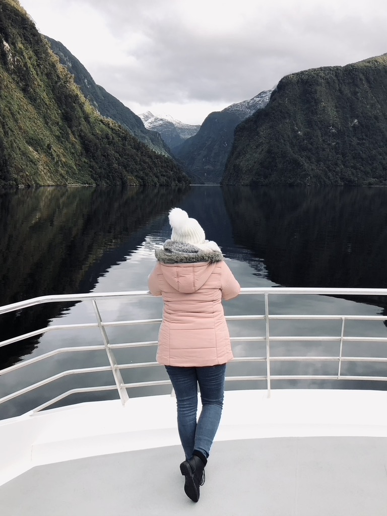 Girl in pink jacket on boat tour with green mountains and water reflection at Doubtful Sound, New Zealand
