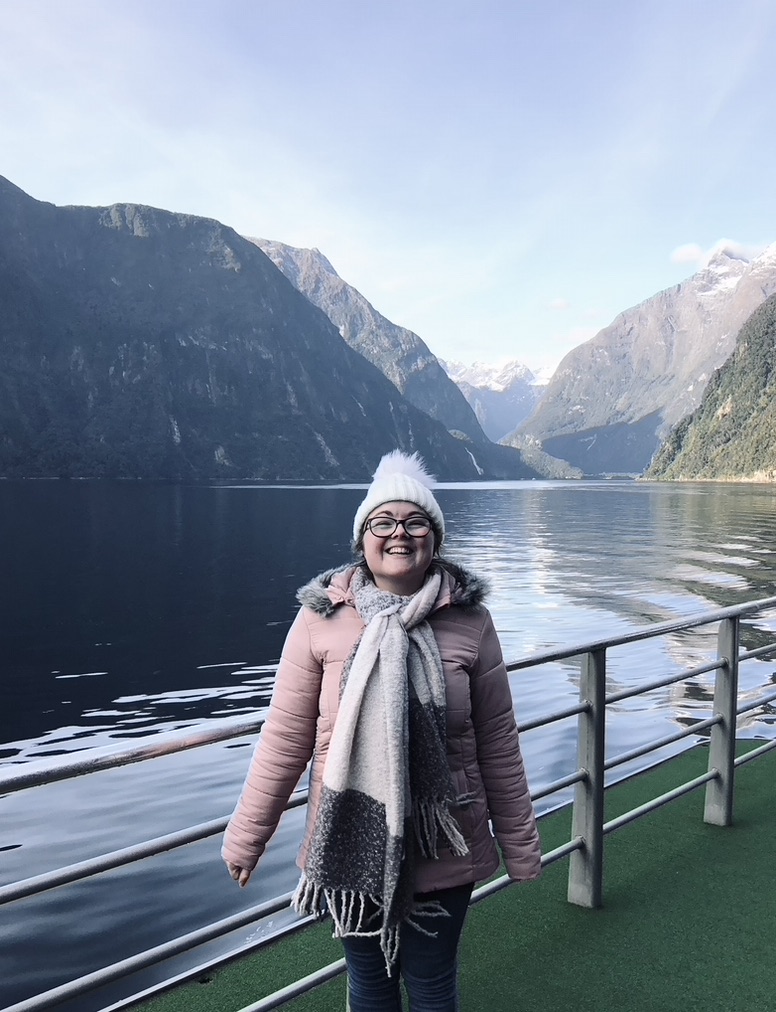 Girl in pink jacket on boat tour with green mountains and water reflection at Milford Sound, New Zealand