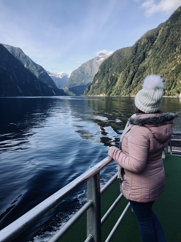 Girl in pink jacket looking out at fjords on on boat tour with green mountains and water reflection at Milford Sound, New Zealand