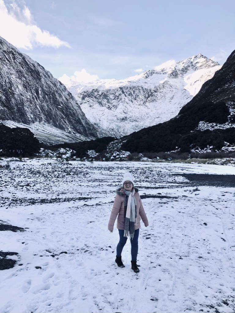 Girl in pink jacked walking in snow with snow capped mountains in the background on road to MIlford Sound in New Zealand