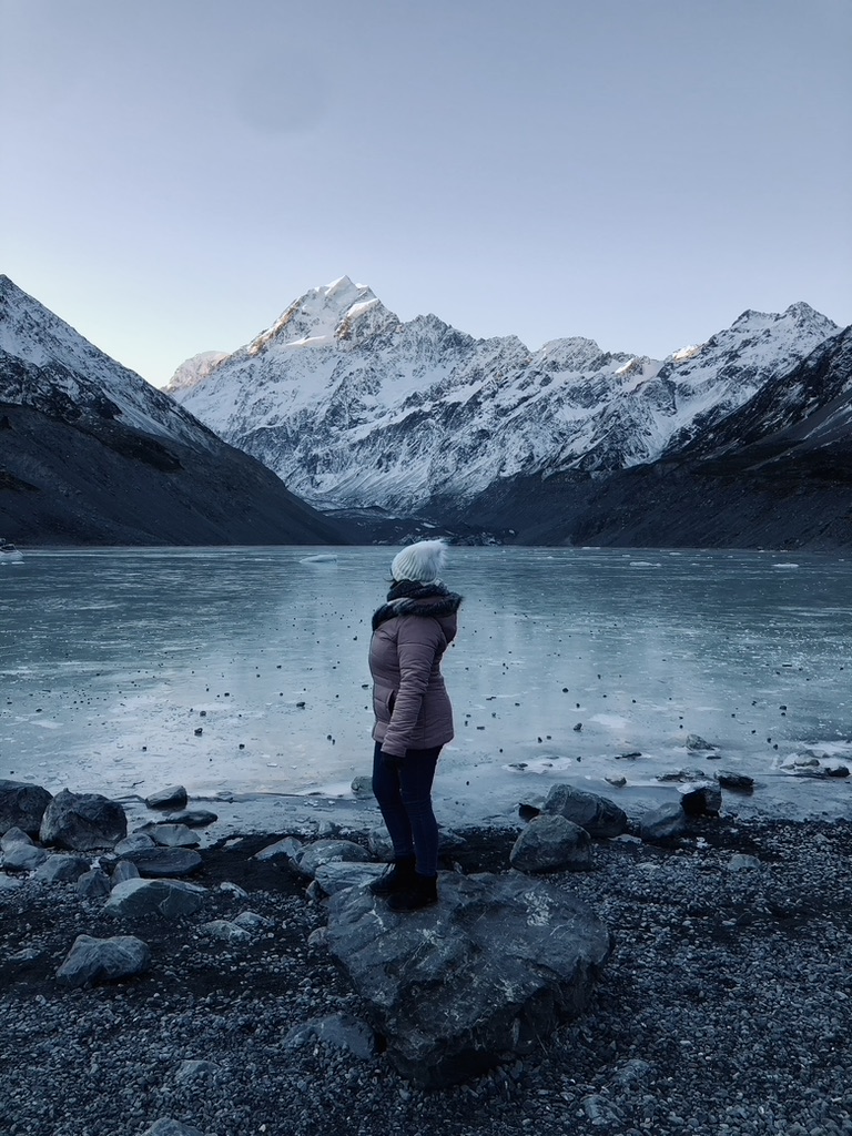Girl in pink coat looking back at frozen lake with Mt Cook on the Hooker Valley Track, New Zealand