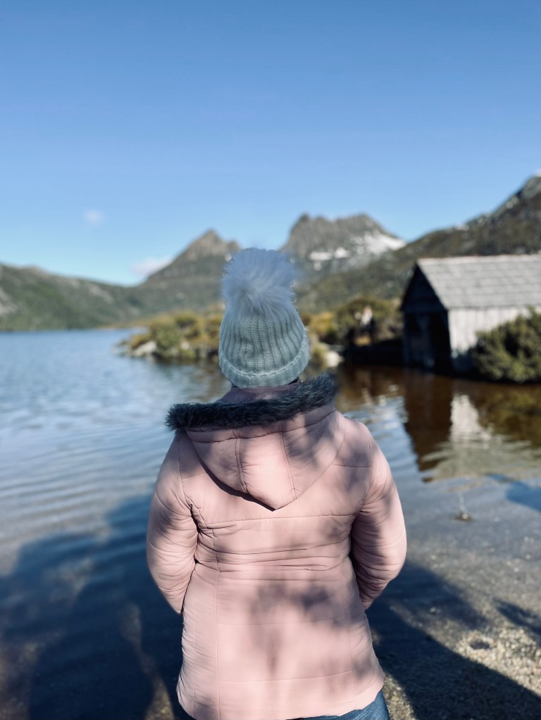 Girl in pink coat looking out at Cradle Mountain with lake and old wooden house in Tasmania, Australia